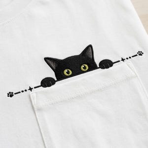 May include: A white t-shirt featuring an embroidered design of a black cat peeking over the top edge of a chest pocket. The cat has bright yellow eyes and is shown with its front paws resting on the pocket edge, accented by a horizontal line.