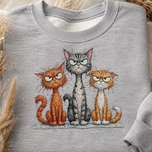 May include: A heather gray crewneck sweatshirt featuring a graphic illustration of three wet, grumpy-looking cats sitting together. The cats are depicted in orange and gray tabby patterns with furrowed brows and ruffled fur, appearing damp.