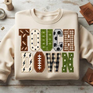 Könnte beinhalten: Ein cremefarbenes Sweatshirt mit Rundhalsausschnitt und dem Wort TOUCHDOWN in großen Blockbuchstaben. Jeder Buchstabe hat ein einzigartiges Muster, darunter Footballs, Sterne, ein Spielfeldraster, Helme, braunes Karomuster und schwarz-weißes Karo.