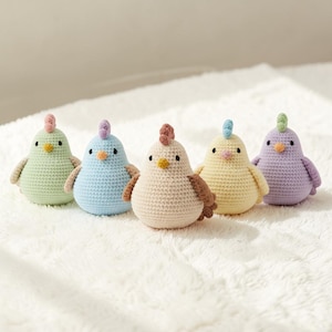 May include: Five handmade crochet chicks in pastel colors: light green, blue, beige, yellow, and purple. Each chick has a different colored comb and beak. The chicks have small wings and are sitting on a white, textured surface.