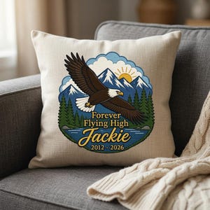 May include: A square off-white throw pillow featuring a circular graphic of a bald eagle in flight over a lake, pine trees, and snow-capped mountains with a sun. The text reads Forever Flying High, the name Jackie, and the years 2012-2026.