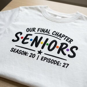 May include: A white t-shirt featuring black embroidered text that reads "OUR FINAL CHAPTER" above the word "SENIORS" in a bold font. The letters in "SENIORS" are separated by small, colorful embroidered dots. Below is "SEASON: 20 | EPISODE: 27".
