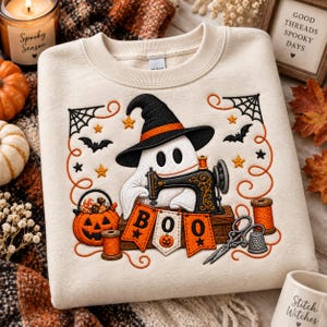 May include: A cream-colored sweatshirt featuring an embroidered design of a ghost wearing a witch hat, operating a sewing machine. The scene includes a jack-o'-lantern bucket, orange thread spools, scissors, a thimble, and banners spelling BOO.