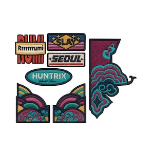 May include: A collection of embroidered patches in various shapes and designs. The patches feature text like "SLAY" and "SEOUL", along with colorful graphics in shades of pink, teal, and gold. The patches are likely for clothing or accessories.