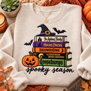 May include: A beige crewneck sweatshirt featuring an embroidered stack of five colorful books topped with a black witch hat. The book spines are labeled The Addams Family, Hocus Pocus, Halloweentown, The Nightmare Before Christmas, and Goosebumps.