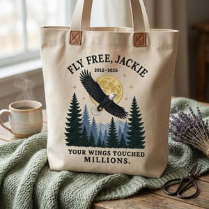 May include: A beige canvas tote bag featuring a graphic of a bald eagle flying over a forest and a full moon. The text reads: FLY FREE, JACKIE 2012-2026 and YOUR WINGS TOUCHED MILLIONS. The bag has two sturdy beige fabric handles with brown accents.