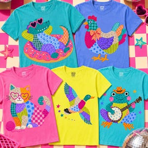 May include: A collection of Comfort Colors t-shirts in various colors, each featuring a whimsical animal graphic. Designs include an alligator in sunglasses, a chicken, a cat, a duck, and a frog playing a banjo. The shirts are displayed on a pink and white checkered surface.