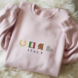 May include: A light pink sweatshirt with embroidered Italian-themed designs. The design includes a gold laurel wreath, the Italian flag, a brown Roman helmet, and the Colosseum. The word "ITALY" is embroidered below the designs.