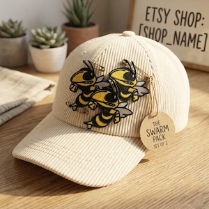 May include: A cream-colored corduroy baseball cap featuring three embroidered yellow and black wasp patches attached with safety pins. A round tan paper tag hangs from the cap, printed with the text THE SWARM PACK SET OF 3.