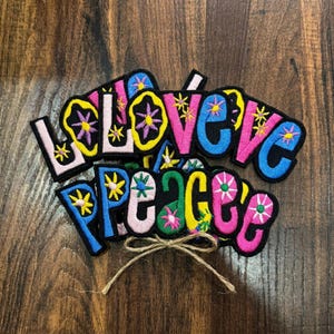 May include: A collection of embroidered fabric patches featuring the words LOVE and PEACE in bold, colorful letters. Each letter is filled with vibrant floral and star patterns in shades of pink, blue, yellow, and purple, outlined in black thread.