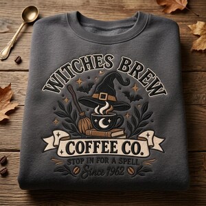 May include: A charcoal grey crewneck sweatshirt featuring a graphic print that reads WITCHES BREW COFFEE CO. STOP IN FOR A SPELL Since 1962. The design includes a witch hat, a broom, a steaming coffee cup with a crescent moon, and bats.