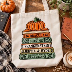 May include: A cream-colored canvas tote bag featuring a graphic print of a stack of five books topped with an orange pumpkin. The book titles are Dracula, The Shining, Frankenstein, Jekyll & Hyde, and The Complete Works of Edgar Allan Poe.