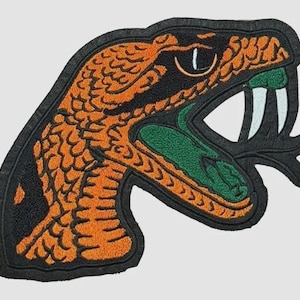 May include: Embroidered patch featuring a snake head with an open mouth. The snake has orange and black scales, a green interior mouth, and white fangs. The patch has a black border and is likely for clothing or accessories.