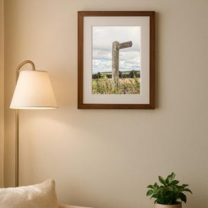 Könnte beinhalten: Gerahmtes Foto eines verwitterten Holzwegweisers auf einem Feld. Der Wegweiser hat einen Richtungsarm und ist vor dem Hintergrund eines bewölkten Himmels und grüner Felder angebracht. Der Rahmen ist dunkelbraun.