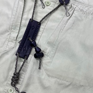 May include: Close-up of a fishing lanyard with a dark blue leather tag and decorative elements. The lanyard is attached to a light green shirt with the brand name "SIMMS" visible. The lanyard features a key ring and various clips.