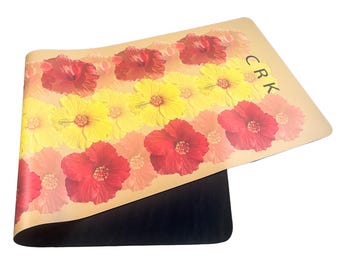 May include: A yoga mat featuring a floral design with red and yellow hibiscus flowers. The flowers are arranged in rows against a tan background. The mat has a black underside and the letters "CRK" are visible.