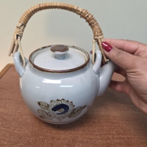 Peut inclure: Théière en céramique bleu clair avec un bord et un couvercle marron. La théière présente un motif de tournesol et une anse tressée. La poignée est en matériau marron clair. La théière est conçue pour infuser du thé.