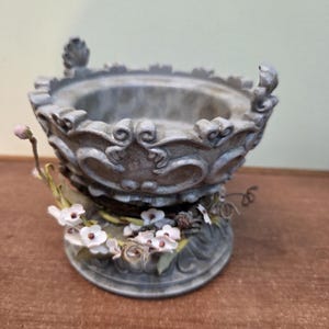May include: An ornamental grey bowl with a floral design. The bowl has a detailed, textured surface and is decorated with small white flowers and green leaves around the base. The rim features decorative scrollwork.