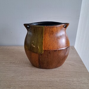 Op de afbeelding: Een houten vaas met een zwarte rand en twee handvatten. De vaas is geschilderd in tinten bruin en groen, waardoor een schaakbordpatroon ontstaat.