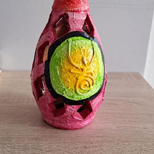 Puede incluir: Un jarrón decorativo con un exterior rosa texturizado y un borde rojo. El jarrón presenta formas geométricas recortadas y un diseño central con acentos verdes, amarillos y negros. El jarrón está sobre una superficie de madera.