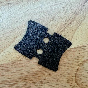May include: A black, textured metal decorative clasp with two circular holes. The clasp has a unique, geometric shape with pointed edges and is set against a light brown wooden surface.
