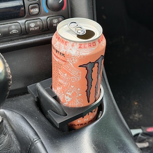 May include: A black plastic cup holder insert holding a peach-colored Monster Ultra energy drink can. The can features white line art graphics, the Monster logo, and text including "Taurine + Zero Sugar" and "Ultra Peachy Keen" on the surface.