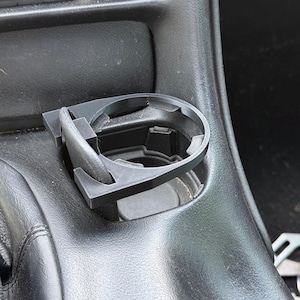 Może przedstawiać: Czarny, wydrukowany w technologii 3D plastikowy uchwyt na kubek, zaprojektowany do montażu w konsoli środkowej pojazdu. Okrągła rama posiada teksturowaną powierzchnię oraz boczny wspornik, który stabilnie mocuje element w otworze konsoli.
