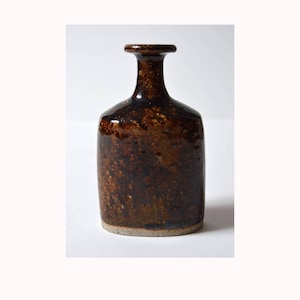 May include: A small, brown ceramic vase with a rounded, rectangular body and a narrow neck. The vase has a speckled, brown glaze with a subtle sheen.