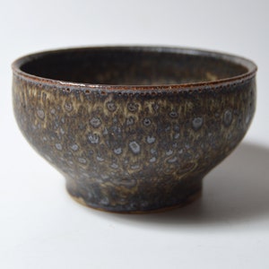 May include: A handmade ceramic bowl with a speckled, textured surface. The bowl features a dark brown exterior with lighter brown and gray accents. The rim has a darker brown trim, and the bowl sits on a small, rounded base.