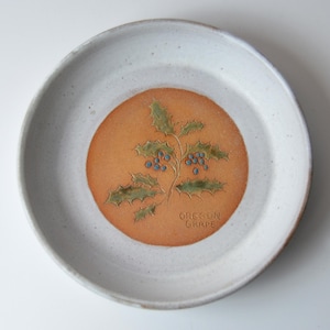 May include: A white ceramic plate with a brown and orange center featuring a green holly sprig with blue berries. The text "Oregon Grape" is printed in the center.