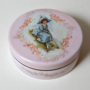 May include: A pink glass trinket box with a white and gold floral design. The lid features a painted image of a girl in a blue dress and a white hat.