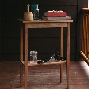 May include: A wooden side table with a shelf. The table has a light brown finish and is made of solid wood. The shelf is made of the same wood and is about 10 inches above the top of the table. There are a few items on the table, including a blue glass jar, a wooden butter mold, and a stack of books. There is also a glass jar and a wood plane on the shelf.