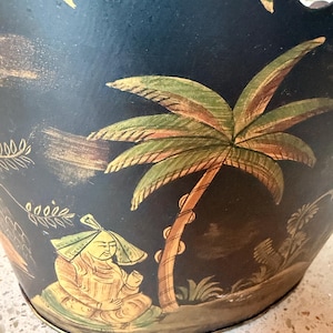 May include: A black metal decorative bowl with a scalloped edge. The bowl features a hand-painted scene with a palm tree, a seated figure, and other tropical elements in shades of gold, green, and brown.