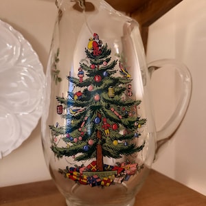 Puede incluir: Una jarra de vidrio transparente con un diseño de árbol de Navidad. El árbol está decorado con adornos, una figura de Papá Noel y regalos envueltos en la base. La jarra tiene un asa y un pico vertedor.