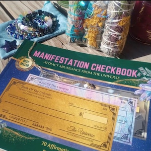 May include: A collection of spiritual items including a "Manifestation Checkbook" with the text "Attract Abundance From The Universe", a gold necklace, bracelets, sage bundles, and a blue star-shaped object. The checkbook has the text "Bank Of The Universe".