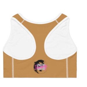 May include: A sports bra in a tan color with white accents and straps. The back of the bra features a racerback design. The bra has the word "ICHI" in a black and pink graphic on the lower back.