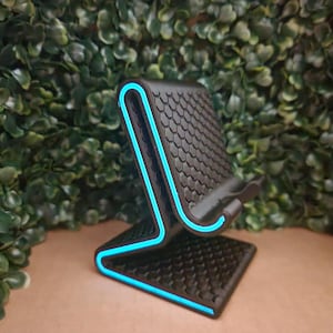 May include: A black phone stand with a textured surface and a neon blue trim. The stand has a unique, modern design with a honeycomb pattern on the phone rest. The stand is designed to hold a phone upright.