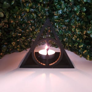 Puede incluir: Portavelas negro en forma de triángulo con un círculo recortado en el centro. Una pequeña vela blanca está ardiendo dentro del portavelas.