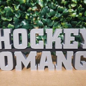 Puede incluir: Un letrero gris impreso en 3D con las palabras "HOCKEY ROMANCE" en mayúsculas. Un palo de hockey negro está posicionado detrás de las letras. El letrero está sobre un fondo de follaje verde.