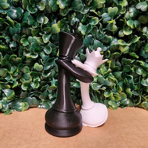 Pode incluir: Uma peça de xadrez preta, em forma de cartola, encosta-se a uma peça de xadrez branca com uma coroa. As peças estão sobre uma superfície castanha com um fundo de folhas verdes. Peças de xadrez decorativas.