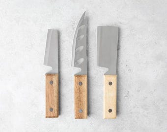 May include: Three cheese knives with wooden handles. The knives have different blade shapes and are arranged in a row on a light gray surface.