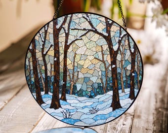 Op de afbeelding: Een rond glas-in-loodraam dat aan een ketting hangt. Het raam toont een winterscène met bomen en sneeuw in tinten blauw, bruin en wit.