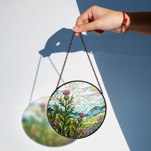 A hand holding a round stained glass piece with a colorful floral design, against a white background.