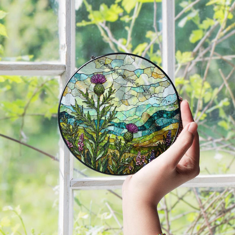 A hand is holding a stained glass piece featuring a vibrant landscape scene with a thistle and other flowers.