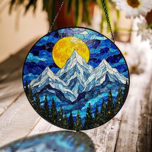 May include: A stained glass window hanging with a depiction of a mountain range with a large yellow sun in the background. The mountains are blue and white with a dark blue sky. The window is round and has a metal frame.