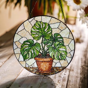 Op de afbeelding: Een hangend glas-in-loodraam met een afbeelding van een monstera-plant in een pot met groene bladeren tegen een witte en gele achtergrond.