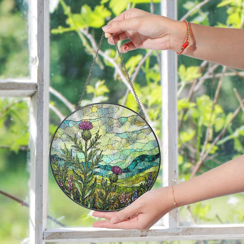 A person's hand holding a stained glass piece with a colorful landscape scene, including a thistle flower, in front of a window with green leaves.