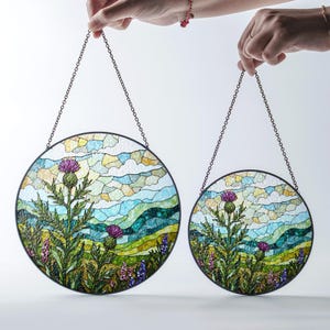 Two hands holding round stained glass panels with a landscape scene featuring a thistle and purple flowers.