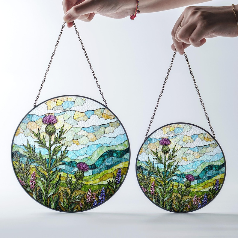 Two hands holding round stained glass panels with a landscape scene featuring a thistle and purple flowers.