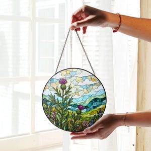 A person's hands holding a round stained glass piece with a colorful landscape scene depicting a thistle and other flowers.